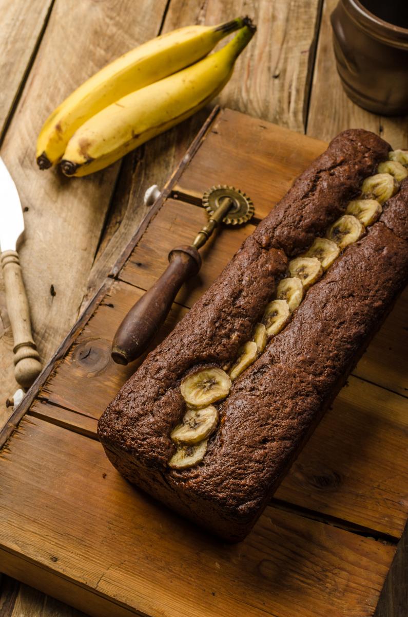 A loaf of banana bread topped with banana slices served on a wooden board.